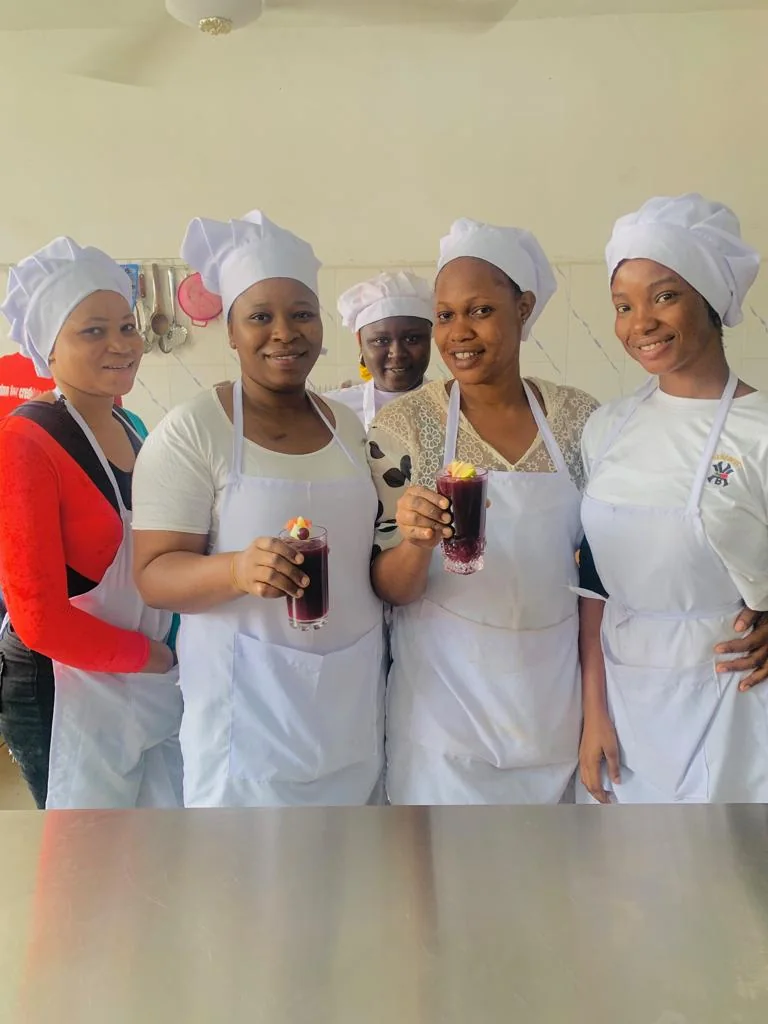 A group wearing aprons and chef hats holds prepared drinks in a training kitchen.