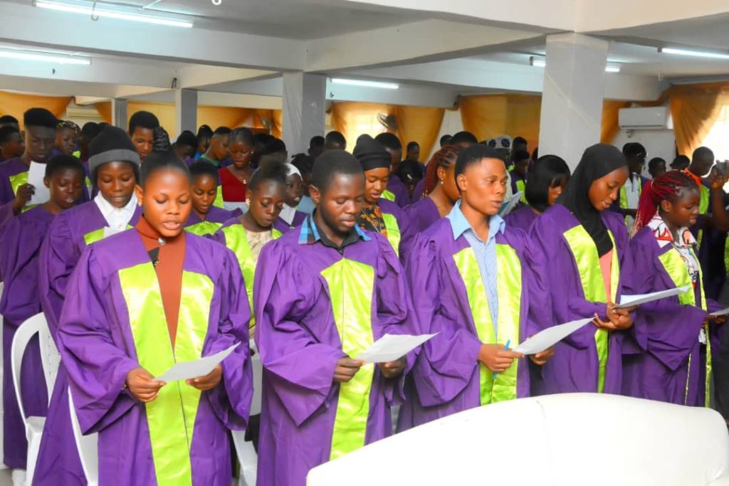 Participants in academic gowns hold printed sheets during an indoor ceremony.