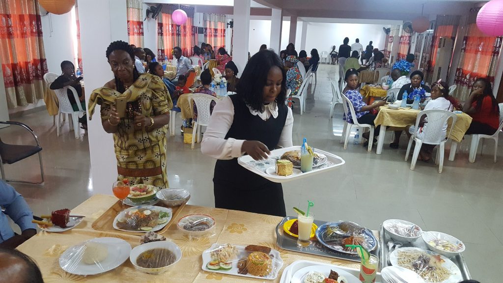 A participant carries a tray of prepared food beside a display table at an indoor gathering.