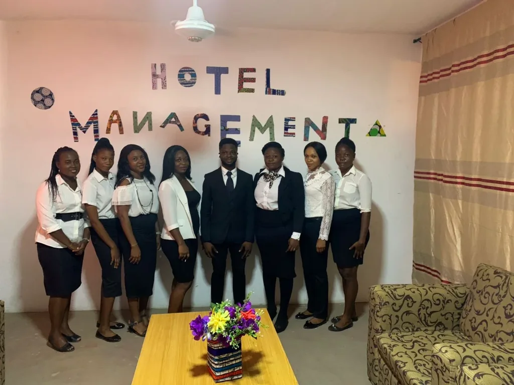 A group in formal clothing stands in a room labelled Hotel Management.