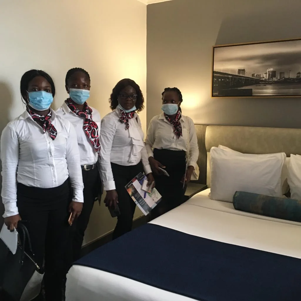 Four visitors in formal clothing stand beside a made bed in a guest room.