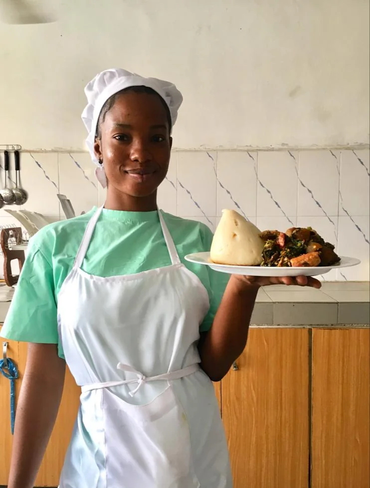 A learner in a chef hat and apron presents a plated meal in a kitchen.
